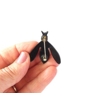 May include: A black bee-shaped brooch with a gold pin backing. The brooch has a simple, stylized design with wings and a segmented body. The pin is held in a hand against a white background.