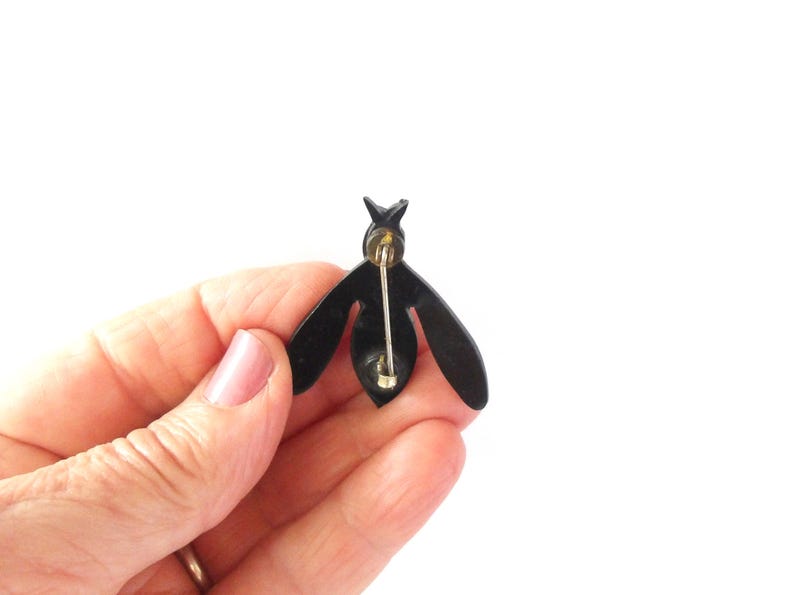 May include: A black bee-shaped brooch with a gold pin backing. The brooch has a simple, stylized design with wings and a segmented body. The pin is held in a hand against a white background.