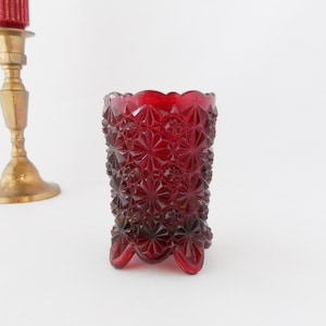 May include: A ruby red, vintage-style glass votive candle holder with a textured, starburst pattern. The small, footed glass has a scalloped rim. A brass candlestick with a red candle is in the background.