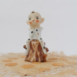 Può includere: Statuina vintage in ceramica di un elfo seduto su un ceppo d'albero. L'elfo ha un incarnato chiaro, guance rosee e pois neri sul suo abito bianco. Il ceppo d'albero è marrone con accenti dorati.