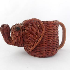 Puede incluir: Una cesta marrón tejida con forma de elefante. La cesta tiene una trompa, orejas y un asa. La cesta de elefante está hecha de material tejido y es un objeto decorativo.