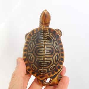 Puede incluir: Figura de tortuga de cerámica con un patrón de caparazón detallado en tonos marrones y beige. La cabeza y las patas de la tortuga son visibles, y tiene un pequeño agujero en la parte inferior. La figura se sostiene en una mano.