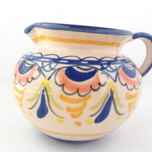 May include: A ceramic jug with a rounded body and a handle. The jug is painted with a cream-coloured base and decorated with blue, orange, and yellow floral patterns. The rim and handle are blue.