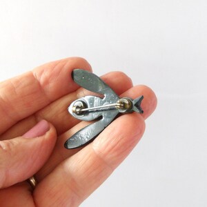 May include: A dark gray metal bee-shaped brooch. The brooch has a pin on the back for attachment. The wings and body are detailed with etched lines. The brooch is held in a hand.