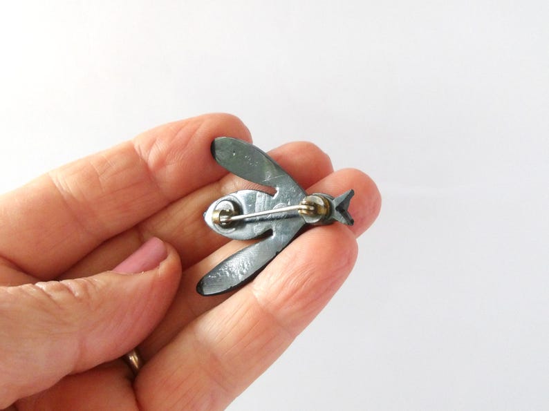 May include: A dark gray metal bee-shaped brooch. The brooch has a pin on the back for attachment. The wings and body are detailed with etched lines. The brooch is held in a hand.