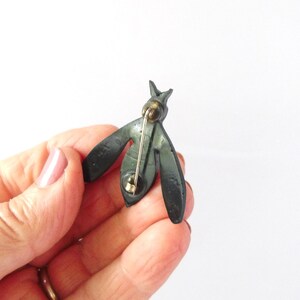 May include: A dark green metal bee-shaped brooch. The brooch has a pin on the back for attachment. The wings are stylized and spread out. The brooch is held in a hand against a white background.