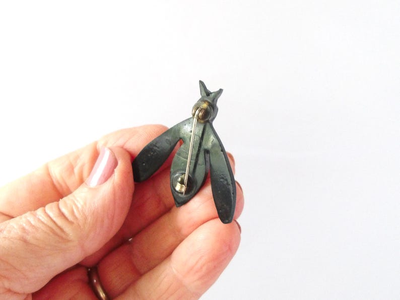 May include: A dark green metal bee-shaped brooch. The brooch has a pin on the back for attachment. The wings are stylized and spread out. The brooch is held in a hand against a white background.