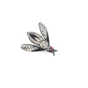 May include: A black and silver insect brooch with three wings. The wings are adorned with clear and red rhinestones. The brooch is set against a white background.