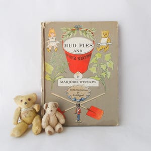 Könnte beinhalten: Ein Vintage-Kinderbuch mit dem Titel "Mud Pies and Other Recipes" von Marjorie Winslow, mit Illustrationen. Zwei kleine Teddybären sitzen vor dem Buch. Das Buchcover zeigt einen roten Eimer und skurrile Illustrationen.