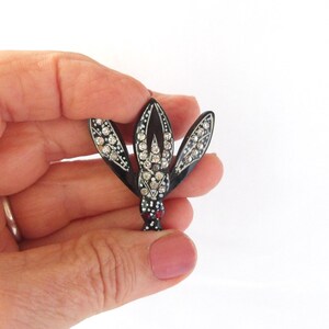 May include: A black and silver brooch in the shape of a stylized flower. The brooch features three petals adorned with clear rhinestones and a central cluster of larger stones. The base has red and white accents.