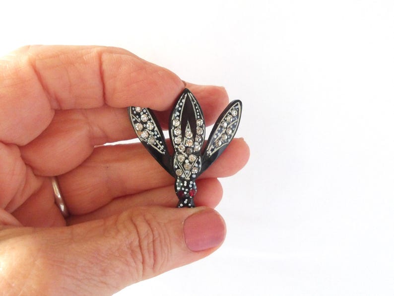 May include: A black and silver brooch in the shape of a stylized flower. The brooch features three petals adorned with clear rhinestones and a central cluster of larger stones. The base has red and white accents.