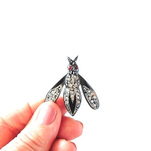 May include: A black and silver insect brooch with rhinestone accents. The brooch has a red stone on the head and is held in a hand against a white background. The wings and body are detailed with sparkling rhinestones.