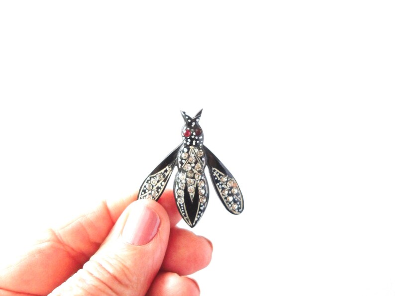 May include: A black and silver insect brooch with rhinestone accents. The brooch has a red stone on the head and is held in a hand against a white background. The wings and body are detailed with sparkling rhinestones.