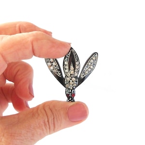 May include: A black and silver brooch in the shape of a stylized flower, held between fingers. The brooch features clear rhinestones and a red accent at the base. The brooch is a vintage accessory.