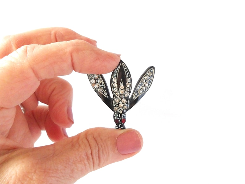 May include: A black and silver brooch in the shape of a stylized flower, held between fingers. The brooch features clear rhinestones and a red accent at the base. The brooch is a vintage accessory.