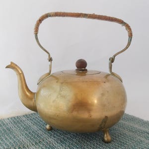 Puede incluir: Una tetera de latón vintage con cuerpo redondeado, boquilla curva y asa trenzada. La tapa presenta un pequeño pomo de madera. La tetera descansa sobre tres pequeños pies. El color general es un dorado cálido y envejecido.