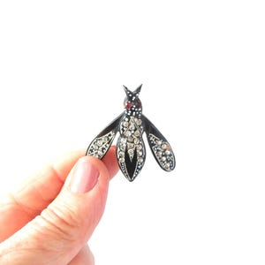 May include: A black and silver insect brooch with a red gem on the head and clear crystals on the wings and body. The brooch is held against a white background.