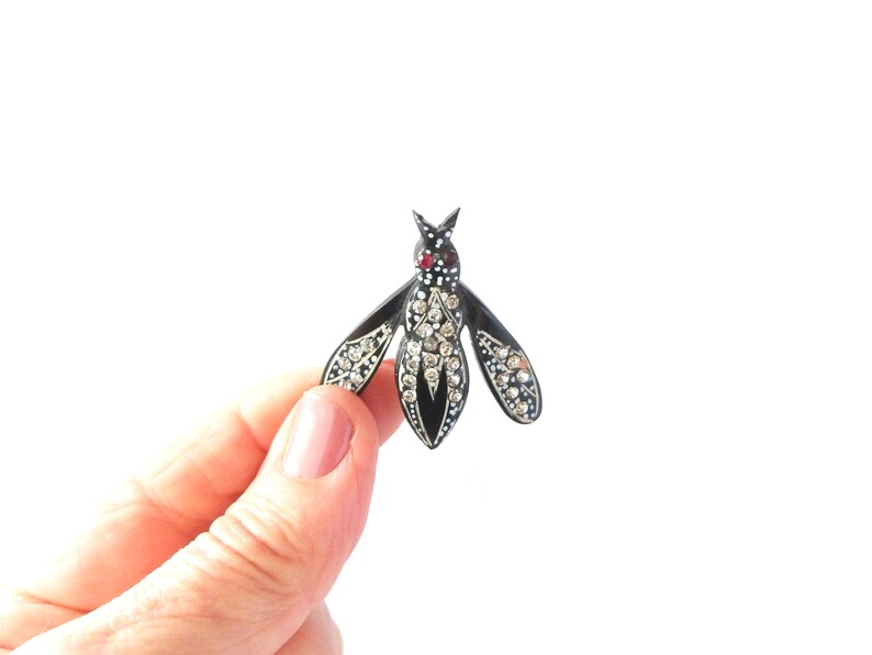 May include: A black and silver insect brooch with a red gem on the head and clear crystals on the wings and body. The brooch is held against a white background.