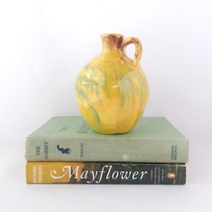 Peut inclure: Une cruche en céramique jaune avec une anse, ornée d'accents verts et bruns. La cruche est posée sur deux livres, un vert et un jaune, avec les titres "The Hobbit" et "Mayflower" visibles.