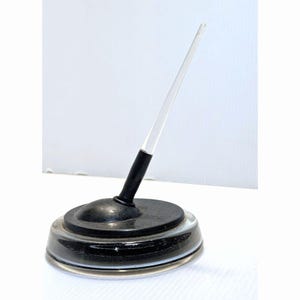 May include: A desk pen set featuring a clear, tapered acrylic pen body with a black grip section. The pen is held upright by a circular black base that sits atop a wider, clear, circular glass or acrylic stand, creating a tiered appearance.