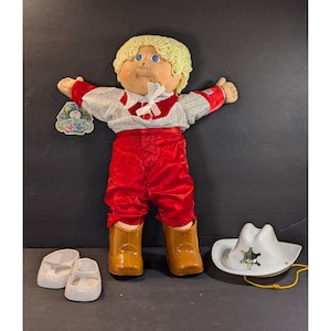 May include: A vintage Cabbage Patch Kid doll in a silver and red outfit, cowboy boots, and a white cowboy hat with a sheriff's badge. Includes white shoes. The doll has blonde hair and blue eyes.