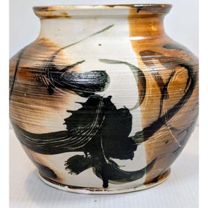 以下が含まれることがあります： 幅広で丸みを帯びた胴体と短い首を持つ陶器製の花瓶。表面はクリーム色のベースに水平な茶色の筆致が施され、容器の外側を包み込むような大胆で黒い抽象的な模様が描かれています。