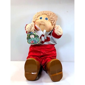 Könnte beinhalten: Eine Cabbage Patch Kids Puppe mit blonden Garnhaaren, blauen Augen und einem rosigen Teint. Die Puppe trägt ein grau-rotes Oberteil, eine rote Hose und braune Schuhe. Ein Anhänger mit dem Cabbage Patch Kids Logo hängt am Hals der Puppe.