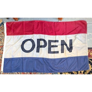 May include: A rectangular horizontal flag featuring three equal horizontal stripes in red, white, and blue. The word OPEN is printed in large, bold, dark blue capital letters across the center white stripe. The flag has a white vertical header.