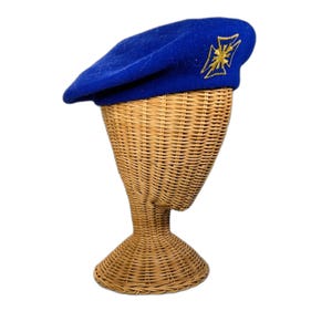 May include: A vibrant royal blue felt beret featuring a gold embroidered cross emblem on the side. The hat has a smooth, rounded crown and a soft texture, showcasing a detailed metallic gold thread design shaped like a cross with a central star.
