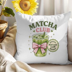 Op de afbeelding: Een wit vierkant kussen met een afbeelding van een groene ijsmatcha-drank in een doorzichtige beker met een roze en witte gingham strik. De tekst luidt MATCHA CLUB en OFFICIAL MATCHA CLUB MEMBER, omringd door kleine groene hartjes en sterren.