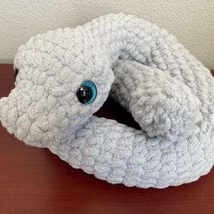 May include: A light gray, plush, crocheted snake toy with large, black eyes and blue accents. The snake is curled up, showcasing its textured, soft body. This handmade item is a cuddly, decorative piece.