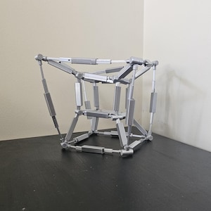 May include: A silver geometric sculpture with a complex, interconnected design. The structure is composed of numerous rods and joints, forming a cube-like shape with open spaces. It sits on a dark surface against a neutral background.