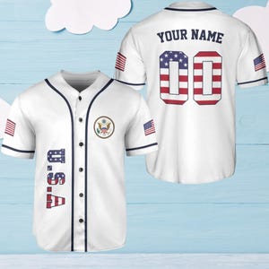 Può includere: Maglia da baseball bianca con finiture blu navy. Il davanti mostra "U.S.A" in rosso e blu, e uno stemma. Il retro presenta "YOUR NAME" sopra il numero "00" in un motivo a stelle e strisce. Toppe della bandiera americana sulle maniche.