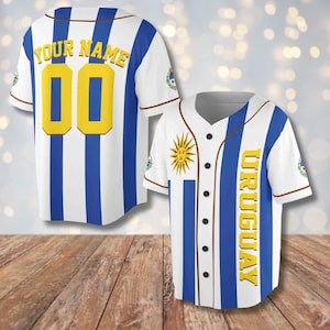 Op de afbeelding: Twee witte honkbalshirts met blauwe verticale strepen en het woord "URUGUAY" in geel. Eén shirt heeft de tekst "YOUR NAME" en het nummer "00" in geel. Het andere shirt heeft een zonnegrafiek.
