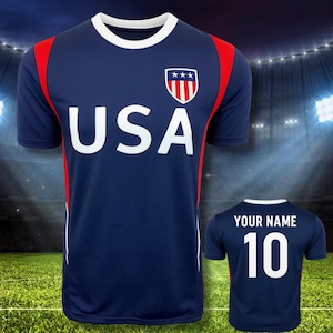 May include: Navy blue USA football shirt with white "USA" text and a shield logo. The shirt has red shoulder accents and a white collar. The back displays "YOUR NAME" and the number "10" in white.