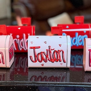 May include: A collection of small, personalized mailboxes in various colors. The mailboxes are primarily pink and red, with names written on them in a cursive font. Each mailbox has a small red flag.