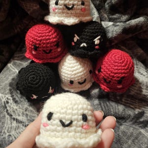 May include: A collection of handmade crochet ghost figures in various colors. The ghosts are white, red, and black, each with a unique facial expression. The crochet ghosts have black eyes and mouths, with pink cheeks. The figures are small and round.