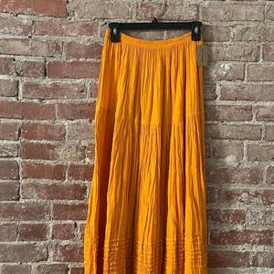 Peut inclure: Longue jupe orange vif suspendue à un cintre noir. La jupe présente un design à plusieurs niveaux avec des détails texturés. Le tissu semble léger. La jupe est exposée contre un mur de briques rustique. Idéale pour la saison chaude.