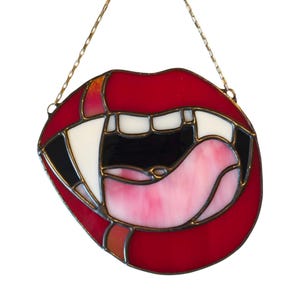 May include: A stained glass artwork showing red lips with white teeth, a black interior, and a pink tongue. The piece is outlined in black and suspended by a gold chain. The lips are slightly parted, creating a striking visual.