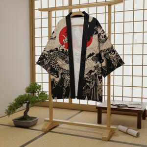 Puede incluir: Kimono beige con un diseño de dragón en blanco y negro y un sol rojo. El kimono está colgado en un estante de madera frente a una pantalla japonesa tradicional. Las mangas son anchas y la tela tiene dibujos.