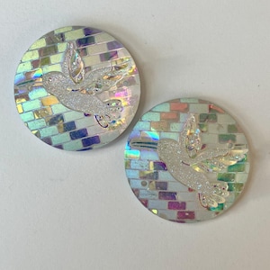 May include: Two round mosaic coasters with a shimmering, iridescent finish. Each coaster features a white, glittery hummingbird design. The mosaic background has a brick-like pattern with a mix of pastel colors.