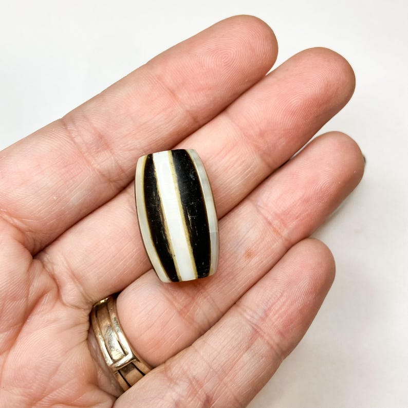 May include: A barrel-shaped bead with a black and white striped pattern. The bead has a polished finish and is held in an open hand. The stripes are vertical and evenly spaced, creating a bold contrast.