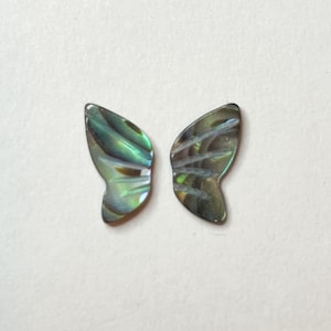 May include: Two iridescent abalone shell butterfly wing shapes. The wings display a range of colors, including green, blue, and hints of purple, with a textured, pearlescent surface. The wings are likely for jewelry making or craft projects.