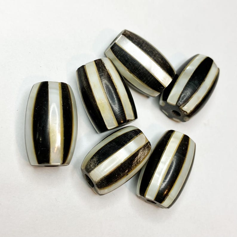 May include: A collection of six barrel-shaped beads. Each bead features alternating black and white stripes. The beads have a glossy finish and are arranged on a white surface. The beads are likely for jewelry making.