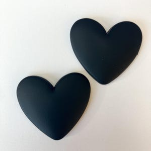 May include: Two matte black heart-shaped objects are displayed against a white background. The hearts have a smooth, rounded appearance and are positioned at a slight angle to each other. The objects are likely decorative or functional.