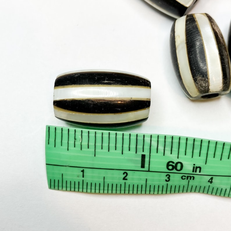May include: Close-up of several black and white striped beads. The beads are oval-shaped and have a glossy finish. A green ruler is placed next to the beads, showing measurements in inches and centimeters.