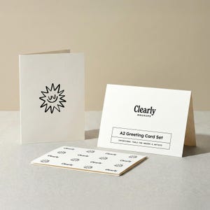 May include: A set of A2 greeting cards in a neutral color. One card features a smiling sun design, while another displays the text "Clearly Mockups" and "A2 Greeting Card Set". A third card shows a repeating pattern of the "Clearly" logo.