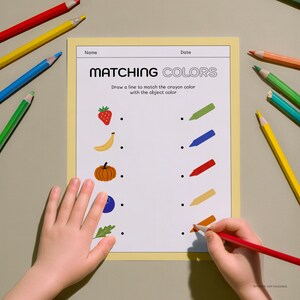 May include: A child's hands are shown working on a "Matching Colors" worksheet. The worksheet features illustrations of a strawberry, banana, pumpkin, blueberry, and leaf. Colored pencils in red, yellow, blue, and green surround the worksheet.