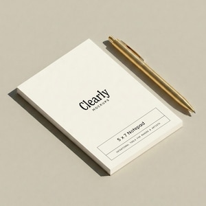 May include: A 5 x 7 inch notepad with the text "Clearly Mockups" and "5 x 7 Notepad" in black. A gold pen rests on the notepad. The notepad is white and the pen is gold.