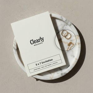 May include: A mockup of a 5 x 7 invitation with the text "Clearly Mockups" and "5 x 7 Invitation". The invitation is on a marble tray with two gold rings and a gold necklace.